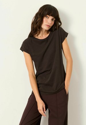 Woman with long brown hair wearing a dark brown short-sleeve shirt and matching pants, standing against a plain light background.