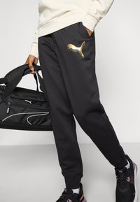 Sorte joggebukser med et gull Puma-logo, sidelommer og elastiske mansjetter, kombinert med en svart treningsbag og svarte sneakers.