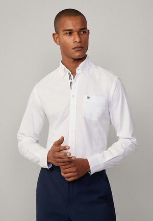 Witte katoenen shirt met een button-down kraag, borstzak en marineblauwe accentueringen op de binnenmanchetten. Model poseert met de handen in elkaar.