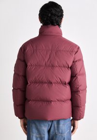 Maroon puffer jacket featuring a high collar, quilted design, and elastic cuffs, paired with blue denim jeans. Smooth fabric texture.