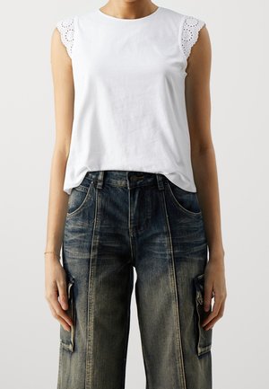 Femme portant un haut blanc sans manches avec des manches en dentelle ajourée et un jean cargo large bleu foncé délavé.