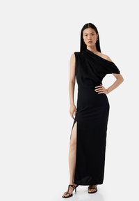 Langes schwarzes Kleid mit einem schulterfreien Design, seitlichem Schlitz und weichem, fließendem Stoff. Kombiniert mit schwarzen Riemchenheels.