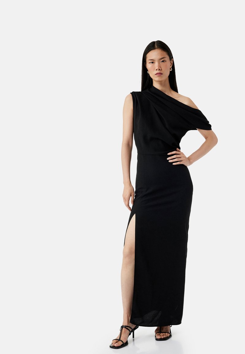 Langes schwarzes Kleid mit einem schulterfreien Design, seitlichem Schlitz und weichem, fließendem Stoff. Kombiniert mit schwarzen Riemchenheels.