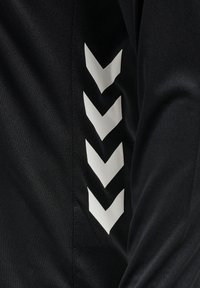 Sort atletisk jakke med hvide chevron-mønstre langs ærmet, lavet af glat, letvægts materiale med en tætsiddende design.