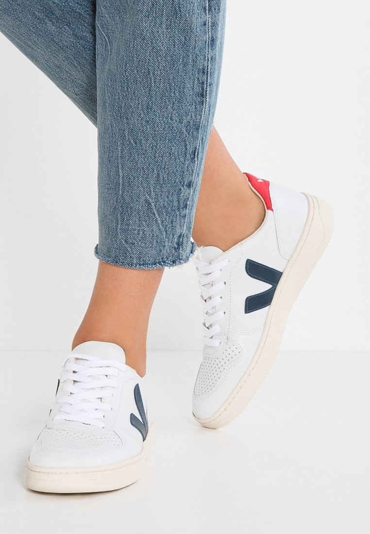 Personne portant des baskets blanches avec un logo "V" bleu marine et un détail rouge au talon, associées à un jean bleu court, debout sur un sol blanc.