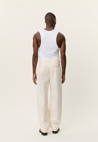 Λευκό μπλουζάκι χωρίς μανίκια, κρεμ παντελόνι με φαρδιά μπατζάκια και υφή, με ορατές ραφές στην πλάτη και τσέπες. Το μοντέλο στέκεται γυρισμένο προς τα πίσω σε ανοιχτό φόντο.