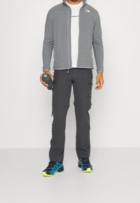 Szary rozpinany kardigan, biały t-shirt, ciemnoszare spodnie z zamkami i czarne/tealowe buty sportowe. Trzyma ciemną butelkę na wodę.
