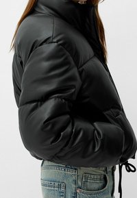 Svart vadderad läderlik jacka som bärs med ljusblå denimjeans, som visar sidovy av torso och arm med handen i fickan.