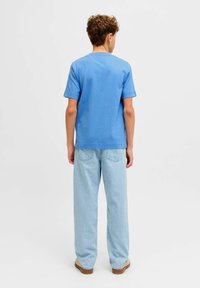 Νεαρό άτομο με κατσαρά μαλλιά στέκεται πλάτη, φοράει μπλε πουκάμισο με κοντά μανίκια, ανοιχτόχρωμο τζιν και μπεζ παπούτσια.