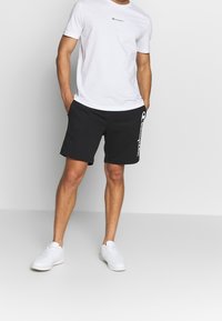 Hvid kortærmet T-shirt med sort logo, parret med sorte shorts med hvidt logo på siden, båret med hvide sneakers.