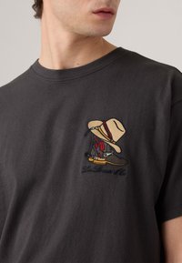 Acercamiento a una persona que lleva una camiseta oscura con un diseño bordado de botas de vaquero y un sombrero en el pecho.