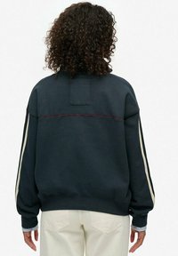 Sweatshirt bleu marine sombre avec une coupe décontractée, présentant des rayures latérales crème, un petit patch au dos et des poignets côtelés. Texture de tissu lisse.