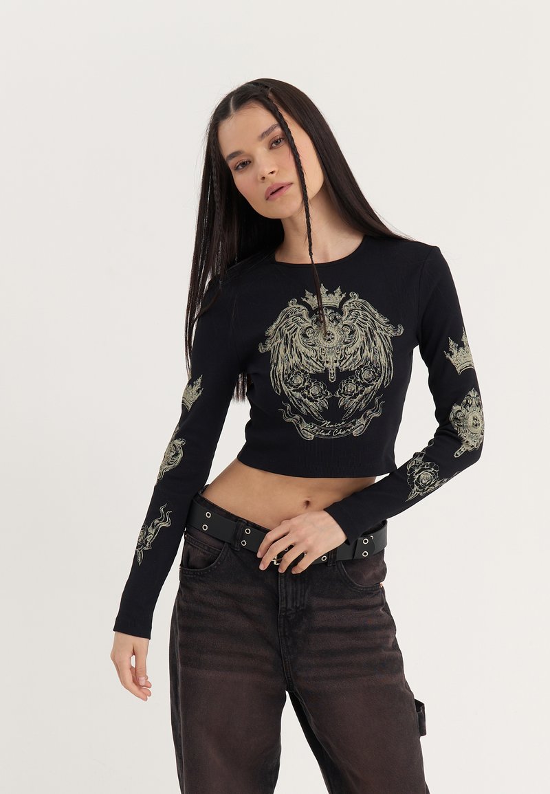 Zwarte lange mouw crop top met gouden leeuw en kroon grafische ontwerpen op de voorkant en de mouwen; geribbelde textuur, getailleerde stijl.
