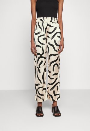 Osoba nosząca kremowe spodnie z wysokim stanem i szerokimi nogawkami z czarnymi abstrakcyjnymi wzorami pędzla oraz czarne sandały na obcasie z odsłoniętymi palcami.