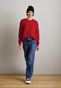 Eine Person geht mit einem roten Pullover, blauen Jeans, einer schwarzen Mütze und schwarz-weißen Turnschuhen auf einem Holzboden vor einer schlichten Wand.