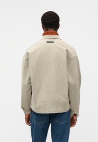 Veste en velours côtelé beige avec un col côtelé orange, une petite étiquette noire à l'arrière, et une coupe décontractée, associée à un jean denim bleu.