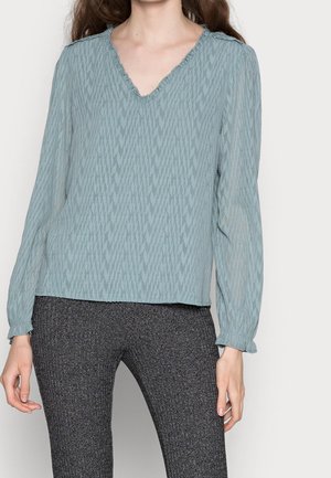 Femme portant un chemisier bleu clair texturé à manches longues avec un col en V à volants et un pantalon texturé gris foncé.