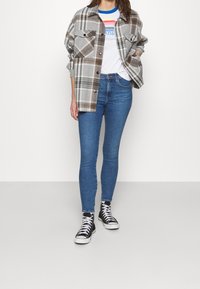 Camisa de cuadros gris con acentos marrones, camiseta gráfica blanca, jeans ajustados azules y zapatillas altas negras. Botones en la camisa y dos bolsillos en el pecho.