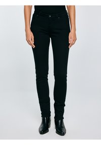 Mujer con vaqueros negros ajustados y botines negros de tacón medio, de pie contra un fondo claro y sencillo.