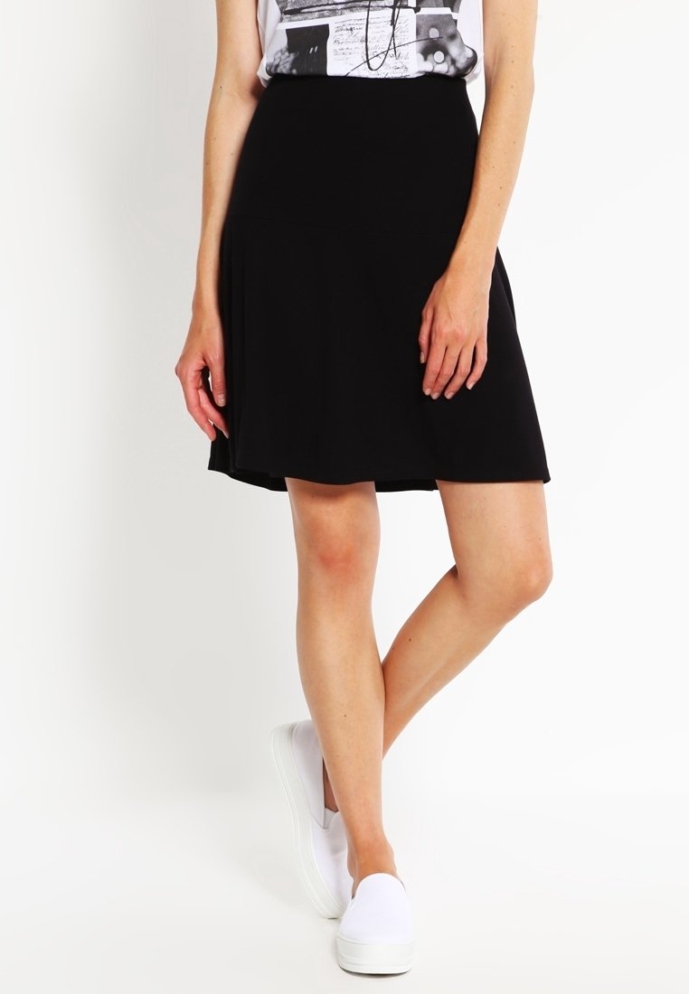 Femme portant une jupe trapèze noire mi-genou, des chaussures blanches sans lacets et un haut sans manches à motif, debout avec les jambes croisées.