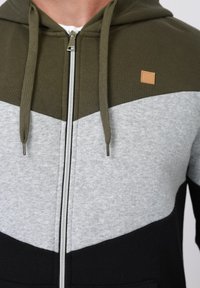 Άνδρας που φοράει φούτερ με φερμουάρ, σε χρώματα χακί, ανοιχτό γκρι και μαύρο, με κορδόνι χακί.