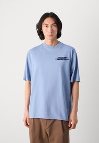 Tricou din bumbac de culoare albastră deschisă, cu guler rotund și mâneci scurte. Prezintă o grafică neagră pe pieptul stâng. Materialul pare moale și neted.