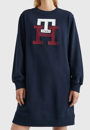 Marineblaues langärmliges Kleid mit großem roten und weißen "TH"-Logo und "Tommy Hilfiger"-Schriftzug vorne, getragen von einer Person vom Hals bis zur Mitte des Oberschenkels.