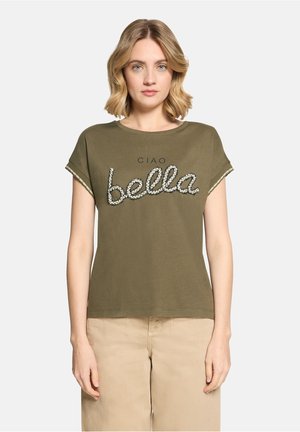 Blonde woman wearing olive green t-shirt with "CIAO bella" text and beige pants, standing against plain light background.