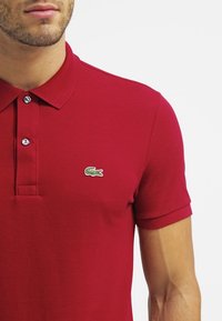 Polo rouge en tissu texturé, à manches courtes, avec une patte de boutonnage à trois boutons et un logo brodé vert sur la poitrine gauche.