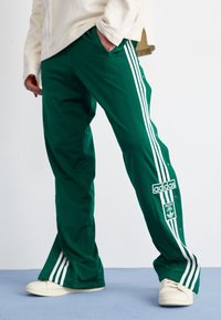 Zelené sportovní kalhoty s bílými postranními pruhy, cvoky a nášivkou adidas. Látka je hladká a má uvolněný střih.