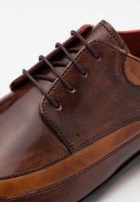 Chaussure en cuir marron avec une finition lisse, dotée de lacets marron, d'un détail cousu le long de la semelle et d'un petit accent rouge à l'intérieur.
