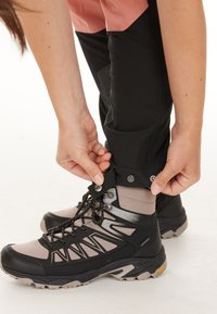Zwarte en grijze wandelschoenen met gestructureerde stof, vetersluiting en verstelbare enkelband. Persoon sluit de band met drukkers.