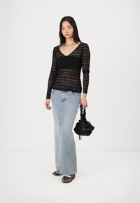 Top lungo in pizzo nero con maniche a sbuffo e scollatura a V, abbinato a una gonna di jeans chiara e scarpe con stampa animale, mentre tiene una borsa nera con coulisse.