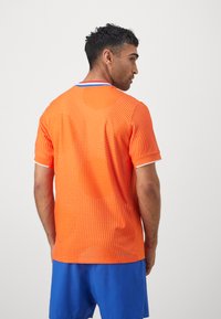 Oranje sportshirt met een stippenpatroon, korte mouwen en een ronde hals met rode en blauwe accenten. Draag met blauwe shorts.