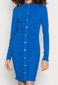 Nainen sinisessä pitkähihaisessa napitettavassa mekossa, jossa on rypytettyjä yksityiskohtia edessä, seisomassa yksiväristä taustaa vasten.