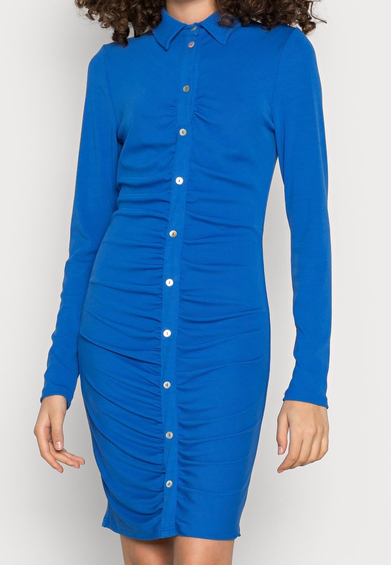 Nainen sinisessä pitkähihaisessa napitettavassa mekossa, jossa on rypytettyjä yksityiskohtia edessä, seisomassa yksiväristä taustaa vasten.