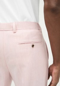 Pantalones ajustados de color rosa claro con una textura suave, que cuentan con un bolsillo trasero asegurado por un pequeño botón marrón. Líneas limpias y diseño de corte slim.