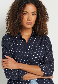 Camisa de botones azul marino en tejido ligero, con patrones de puntos blancos, cuello clásico y mangas enrolladas con cierres de botón.