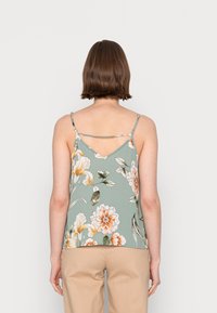 Camisola floreale con spalline sottili, fondo verde, con fiori arancioni e bianchi. Design della schiena a forma di V, tessuto leggero.
