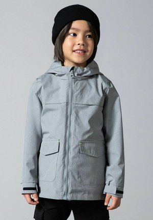 Dziecko ubrane w szarą kurtkę z kapturem i przednimi kieszeniami oraz czarną czapkę, uśmiechające się i lekko patrzące na bok na jednolitym tle.