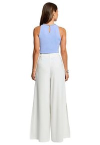 Mujer de espaldas con top azul claro sin mangas con apertura en la espalda y pantalones blancos de pierna ancha y talle alto sobre fondo blanco.