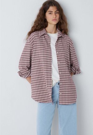 Young woman with long curly hair wears a red and white checkered shirt over a white t-shirt and light blue jeans, standing against a plain wall.