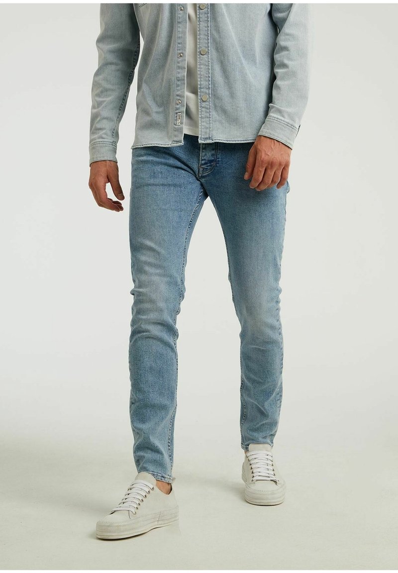 Homme portant un jean slim bleu clair, des baskets blanches et une chemise en jean claire à boutons avec un t-shirt blanc en dessous, debout sur un fond uni.