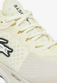 Sneaker Lacoste in crema chiaro, con tomaia in mesh, spesse lace bianche, dettagli testurizzati e una patch con logo nera. Suola LSRFOAM visibile.
