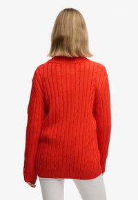 Personne aux cheveux blonds mi-longs portant un pull orange vif torsadé et un pantalon blanc, vue de dos sur un fond uni.