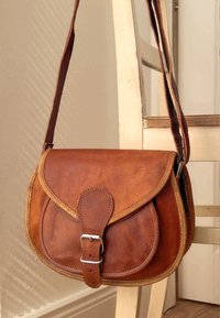 Brązowa skórzana torba na ramię z klapką, metalową klamrą i regulowanym paskiem. Gładka faktura i przeszycia.