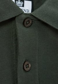 Tmavě zelený strukturální polo límec s dvěma černými knoflíky označenými "Weekend Offender" a částečně viditelnou štítkem.