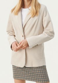 Blazer en velours côtelé beige avec revers à crans et fermeture à un bouton. Associé à une jupe à carreaux aux tons marron, noir et blanc.
