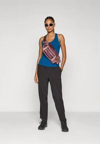 Γυναίκα φορώντας μπλε αμάνικο, μαύρο παντελόνι, μαύρες μπότες, γυαλιά ηλίου και κομψό μαρούν τσαντάκι χιαστί, στέκεται μπροστά σε λευκό φόντο.