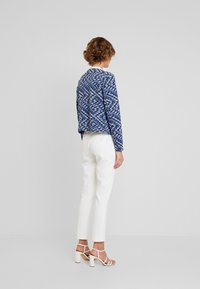 Niebieska kurtka w wzory wykonana z teksturowanej tkaniny, zestawiona z białymi dopasowanymi spodniami i białymi wysokimi obcasami na paski, widziana od tyłu.